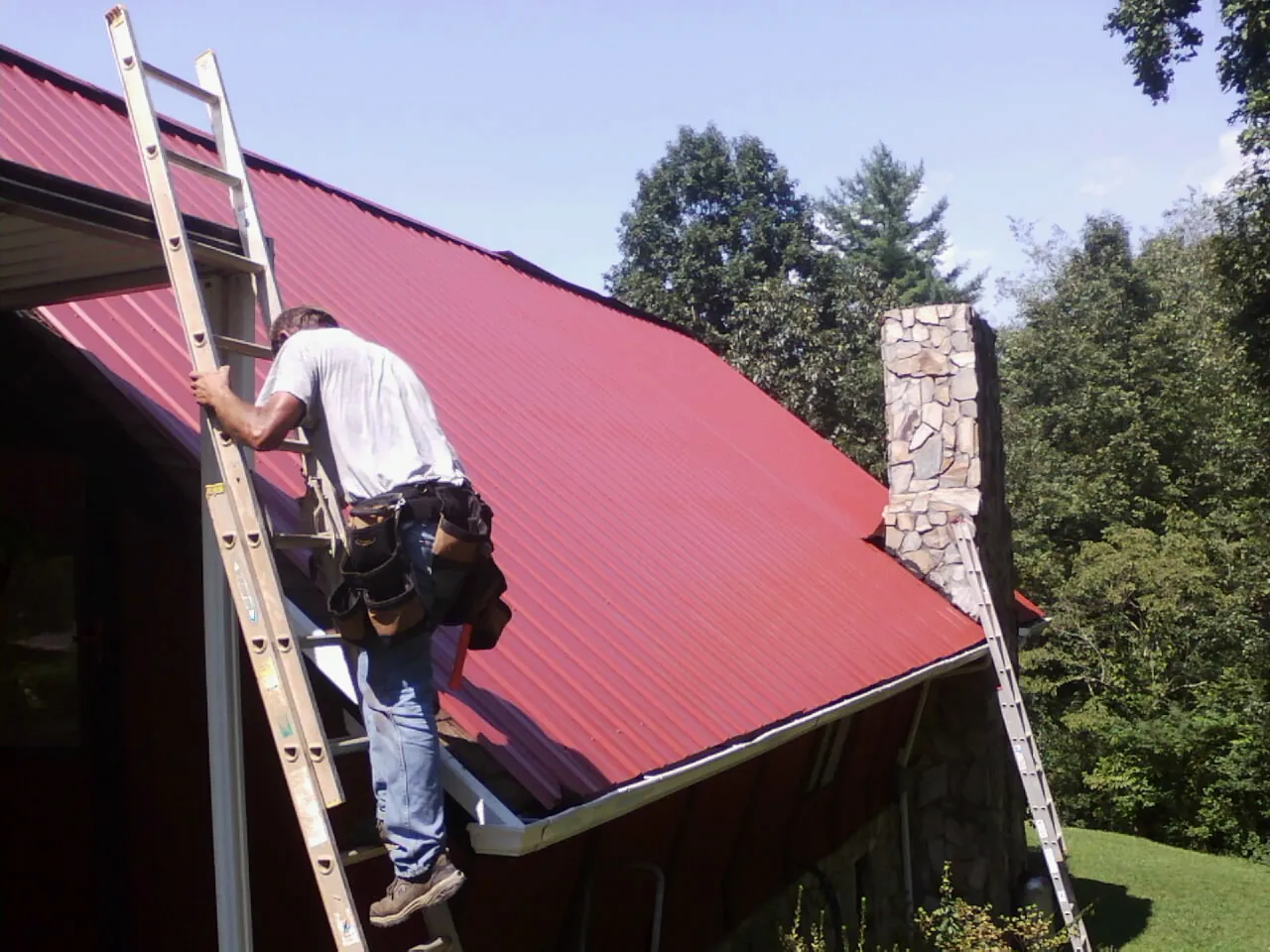 Roofer on a ladder beside a red metal roof and stone chimney — Soffit & Fascia Repair by Roofing Brothers in Turin